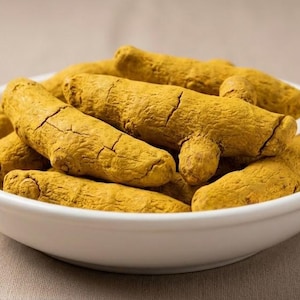 Puede incluir: Un cuenco blanco lleno de raíces de cúrcuma secas. La cúrcuma es de un color amarillo-naranja vibrante, con una superficie rugosa y texturizada y grietas visibles. El cuenco está sobre un fondo beige neutro.