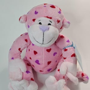 May include: A pink plush monkey toy, decorated with red and purple heart shapes. The monkey has white facial features, hands, and feet. It has a smiling expression and a tag attached to its arm.