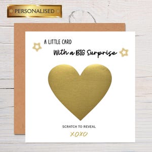 May include: A greeting card with a gold heart-shaped scratch-off area. The card reads "A LITTLE CARD With a BIG Surprise" and "SCRATCH TO REVEAL XOXO". The card is on a brown background and has a gold "PERSONALISED" label.