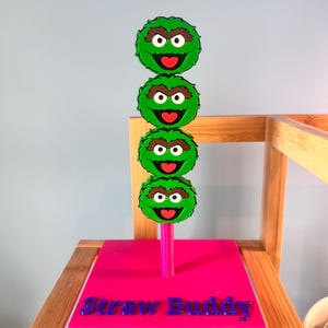 May include: A vertical display stand featuring four stacked green Oscar the Grouch character heads. The stand has a bright pink square base with the text Straw Buddy embossed in blue lettering on the front. The structure is made of plastic.