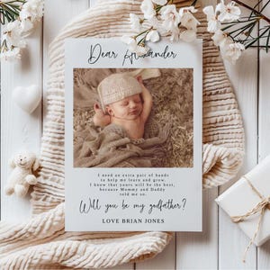 Op de afbeelding: Een geboortekaart met een foto van een slapende baby met een gebreide muts en deken. De kaart bevat de tekst "Dear Alexander" en vraagt "Will you be my godfather?" ondertekend met "Love Brian Jones."