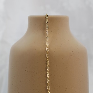 May include: A delicate gold-colored necklace with a floral design hangs against a neutral-toned vase. The necklace features small, repeating flower-shaped links, creating a subtle, elegant aesthetic. The vase has a speckled, matte finish.