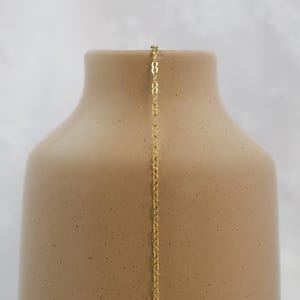 May include: A gold-colored necklace hangs against a beige vase. The necklace has a delicate chain with a repeating pattern. The vase has a smooth, matte finish and a rounded shape. The background is a soft, neutral color.