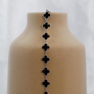 May include: A silver-toned chain bracelet featuring a series of black clover-shaped charms. The bracelet is displayed against a neutral-toned vase, creating a contrast between the jewelry and the background.