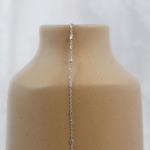 May include: A silver necklace with a delicate chain and small, cylindrical beads. The necklace hangs against a beige ceramic vase, creating a contrast in textures and colors. The necklace is a simple, elegant piece of jewelry.