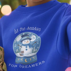 May include: A blue sweatshirt featuring a snow globe design with a snowman, the text "'tis the season" and "FOR DREAMERS". The image is of a smiling child in front of a Christmas tree.