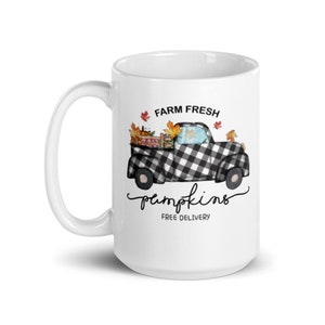 以下が含まれることがあります： 白のセラミックマグ。黒と白のチェック柄のトラックがカボチャと秋の葉を運んでいます。「FARM FRESH pumpkins FREE DELIVERY」の文字がプリントされています。