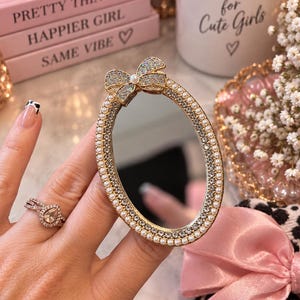 May include: An oval-shaped mirror accessory featuring a gold-toned frame embellished with a border of small white faux pearls and clear crystals. A decorative bow sits at the top, accented with a single central pearl and sparkling crystals.