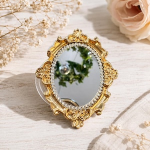 May include: An oval-shaped mirror featuring an ornate gold-colored frame with intricate scrollwork details. The inner edge of the frame is lined with a single row of small, clear, sparkling rhinestones that surround the reflective glass surface.