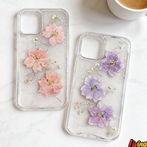 May include: Two clear phone cases with pressed flowers and glitter. One case features pink flowers, the other purple. A white mug of coffee sits nearby. The cases are decorated with small white flowers and glitter.