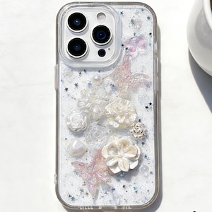 Puede incluir: Funda de teléfono transparente con un diseño decorativo. La funda presenta flores blancas y rosas, mariposas y perlas. La funda tiene un fondo brillante y un borde transparente. La funda está diseñada para un teléfono inteligente.
