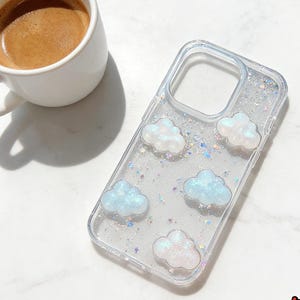 May include: Clear phone case decorated with iridescent glitter and cloud-shaped embellishments in white and light blue. A white coffee cup with espresso sits to the left of the case. The case has a glossy finish.