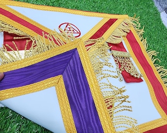 Puede incluir: Un delantal masónico blanco, rojo y morado con flecos y adornos dorados. El delantal presenta una cinta roja, un borde morado y un símbolo en un círculo rojo. El delantal se exhibe sobre una superficie verde.