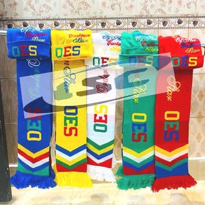 Puede incluir: Una exhibición de coloridas bufandas y gorros de punto. Cada bufanda presenta las letras "OES" y una combinación de colores contrastantes, con gorros a juego encima. Las bufandas tienen extremos con flecos y el texto "Of The Eastern Star".