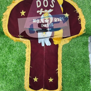 Puede incluir: Una bandera ceremonial burdeos con flecos dorados y detalles de estrellas. La bandera presenta el texto "DOS" y "OASIS" junto con otro texto y un escudo. También se ven un par de guantes blancos.