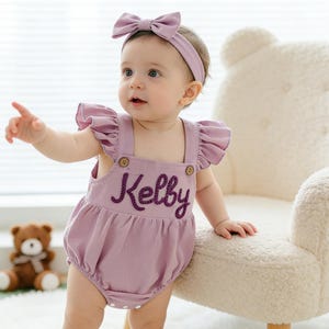 May include: A lavender baby romper with the name "Kelby" embroidered in a darker purple thread. The romper has ruffle sleeves and wooden buttons. A matching headband with a bow completes the outfit.