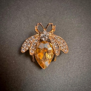 Könnte beinhalten: Goldfarbene Bienenbrosche mit einem großen, facettierten, bernsteinfarbenen Kristall in der Mitte. Die Flügel sind mit kleinen, klaren Kristallen besetzt, und die Brosche hat ein detailliertes Design. Der Körper der Biene ist ein großer, bernsteinfarbener Kristall.