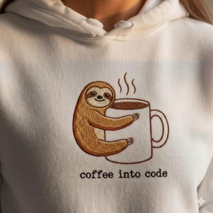 Op de afbeelding: Witte hoodie met een geborduurd ontwerp van een bruine luiaard die een koffiemok omhelst. De mok heeft stoom die eruit komt. De tekst "coffee into code" is eronder geborduurd.