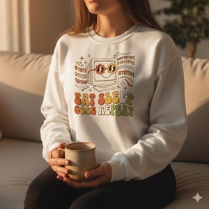 May include: White sweatshirt featuring a retro computer graphic with sunglasses and the text "EAT SLEEP CODE REPEAT" in orange, yellow, and green. The sweatshirt has a crew neck and long sleeves.