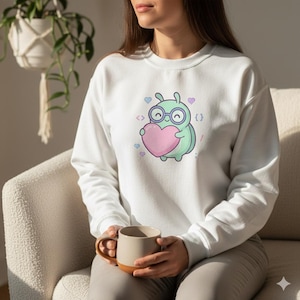 Può includere: Felpa bianca con un personaggio a fumetti a forma di insetto con gli occhiali, che tiene in mano un cuore rosa. L'insetto è verde chiaro con accenti blu. Piccoli cuori e simboli di codice circondano l'insetto. Viene tenuta una tazza con esterno beige e base marrone.