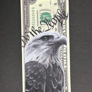 May include: A two-dollar bill with a hand-painted eagle portrait. The words "We the People" are written in black ink across the bill. The bill's details, including the serial number and "Two Dollars" are visible.