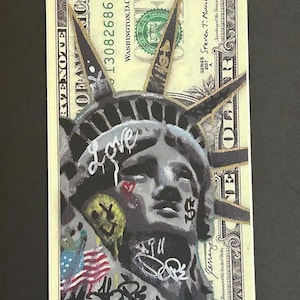Puede incluir: Un billete de un dólar con arte de graffiti. La Estatua de la Libertad está pintada en el billete, con la palabra "Love" y otros grafitis. El billete también presenta una pequeña bandera y la palabra "RISE" en azul.