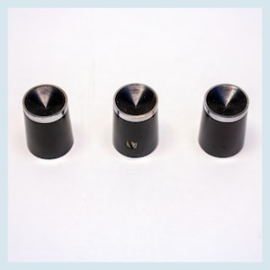 May include: Three black cylindrical knobs with a silver trim and a conical, metallic top. The knobs are evenly spaced against a white background. The knobs appear to be made of metal and plastic.