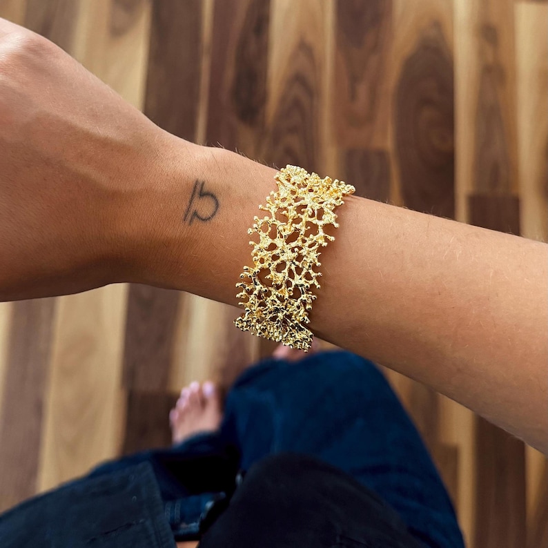 May include: A gold-colored bracelet with a textured, openwork design resembling coral. The bracelet is worn on a wrist with a small tattoo. The background is a wooden floor.