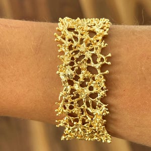 May include: A gold-colored bracelet with an intricate, organic design resembling coral or a network of branches. The bracelet is worn on a wrist and has a textured surface with many small openings. The jewelry piece is a statement accessory.