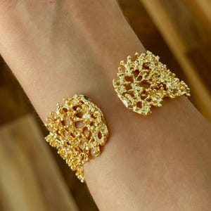 May include: A gold-colored cuff bracelet with an openwork, coral-like design. The bracelet has an organic, textured appearance, with irregular shapes and a bright, reflective surface. The cuff is worn on the wrist, showcasing its intricate details and bold style.