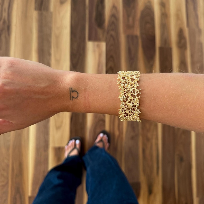 May include: A gold-colored bracelet with a textured, openwork design is worn on a wrist. The bracelet has a wide band and a detailed, organic pattern. A small tattoo is visible on the wrist. The background is a wooden floor.