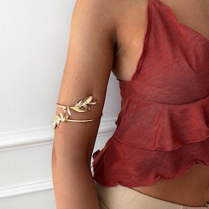 May include: Gold-toned arm cuff featuring a leaf design. The cuff wraps around the upper arm with two bands, adorned with detailed leaf embellishments. The jewelry is shown on an arm, paired with a red top.