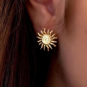 May include: Gold-colored sunburst stud earrings. The earrings feature a textured, hammered finish on the central disc and radiating rays. The earrings are designed to be worn on the ear.