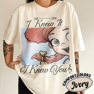 May include: An ivory short-sleeve t-shirt featuring a graphic print of a cartoon character with red hair and a braid. The text EST 2026 I Knew It and I Knew You is printed in black script above and below the image. A small circular label reads Comfort Colors Ivory.