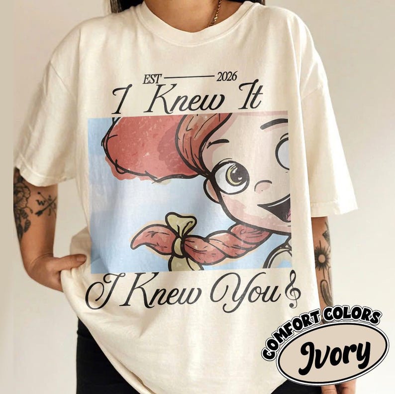 May include: An ivory short-sleeve t-shirt featuring a graphic print of a cartoon character with red hair and a braid. The text EST 2026 I Knew It and I Knew You is printed in black script above and below the image. A small circular label reads Comfort Colors Ivory.