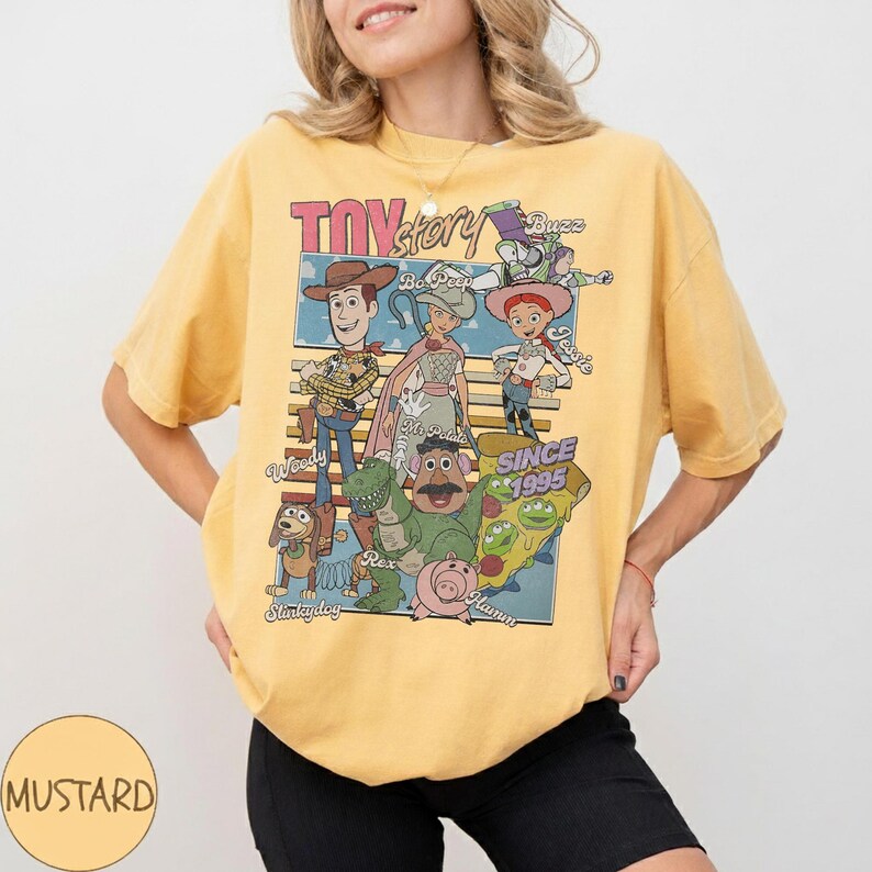 Pu&ograve; includere: T-shirt giallo senape con una grafica dei personaggi di Toy Story, tra cui Woody, Buzz Lightyear e Jessie. Il design include il testo "Toy Story" e "Since 1995". La maglietta ha un girocollo e maniche corte.