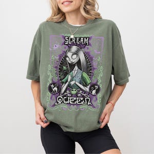 K&ouml;nnte beinhalten: Olivgr&uuml;nes T-Shirt mit einer Grafik einer Figur mit langen wei&szlig;en Haaren, die ein gr&uuml;nes Kleid tr&auml;gt und eine Blume h&auml;lt. Die Grafik enth&auml;lt den Text "SCREAM QUEEN" in Lila und Gr&uuml;n, mit Spinnweben und Katzensilhouetten.