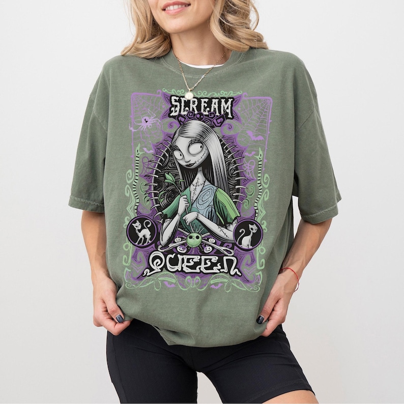 K&ouml;nnte beinhalten: Olivgr&uuml;nes T-Shirt mit einer Grafik einer Figur mit langen wei&szlig;en Haaren, die ein gr&uuml;nes Kleid tr&auml;gt und eine Blume h&auml;lt. Die Grafik enth&auml;lt den Text "SCREAM QUEEN" in Lila und Gr&uuml;n, mit Spinnweben und Katzensilhouetten.
