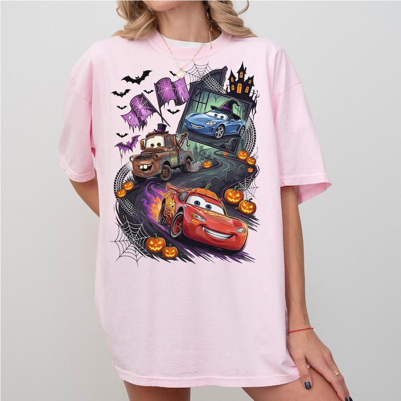 K&ouml;nnte beinhalten: Ein hellrosa T-Shirt mit einem Grafikdruck von zwei Cartoon-Autos, einem braunen Abschleppwagen und einem roten Rennwagen, die auf einer Stra&szlig;e fahren, umgeben von orangefarbenen K&uuml;rbissen, Spinnenweben, Flederm&auml;usen und einem dunklen Spukhaus im Hintergrund.