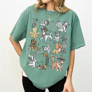 May include: A sage green t-shirt featuring a collection of cartoon dog illustrations. The dogs are in various poses and styles, with hearts and floral accents. The shirt has a relaxed fit and short sleeves.