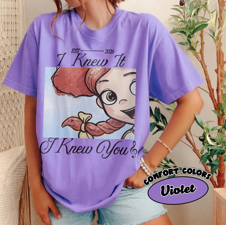 May include: A violet short-sleeved t-shirt featuring a graphic of a cartoon character with red hair in braids. The text above the graphic reads EST 2026 I Knew It, and the text below the graphic reads I Knew You. The shirt has a crew neckline.