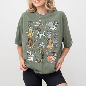 May include: Olive green t-shirt featuring a collection of cartoon dogs in various poses. The dogs are in different colors and breeds, surrounded by hearts and stars. The shirt has short sleeves and a relaxed fit.