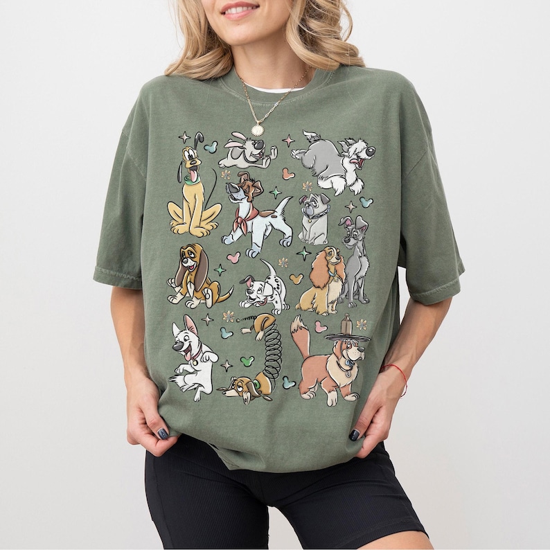 May include: Olive green t-shirt featuring a collection of cartoon dogs in various poses. The dogs are in different colors and breeds, surrounded by hearts and stars. The shirt has short sleeves and a relaxed fit.