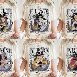 May include: A cream-coloured t-shirt featuring six distinct graphic designs of animated characters wearing witch hats. Each design includes a character name, such as Belle, Elsa, Cinderella, Jasmine, Aurora, or Ariel, surrounded by pumpkins, ghosts, and thorny vines.