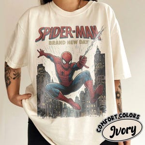 May include: An ivory short-sleeve t-shirt featuring a graphic print of Spider-Man swinging through a city skyline. The text "SPIDER-MAN" and "BRAND NEW DAY" appear above the character. The design includes red and blue colors on the figure.