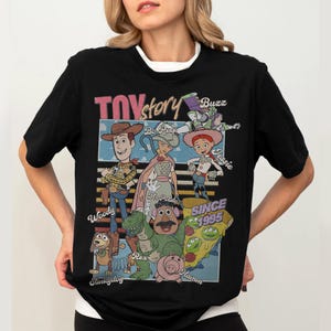 Pu&ograve; includere: T-shirt nera con una grafica dei personaggi di Toy Story, tra cui Woody, Buzz Lightyear e Jessie. Il design include il testo "Toy Story" e "Since 1995". La maglietta ha un colletto bianco.