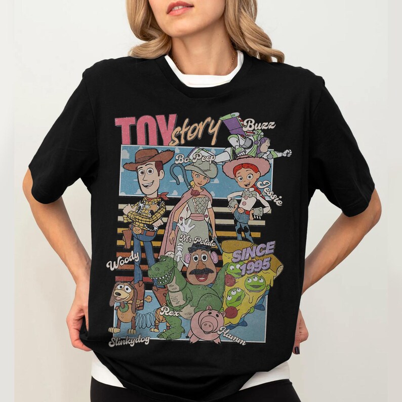 Pu&ograve; includere: T-shirt nera con una grafica dei personaggi di Toy Story, tra cui Woody, Buzz Lightyear e Jessie. Il design include il testo "Toy Story" e "Since 1995". La maglietta ha un colletto bianco.