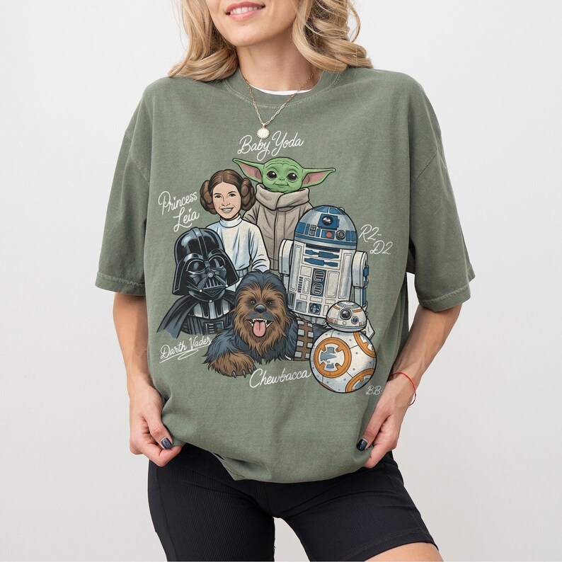 May include: An olive green t-shirt with a graphic of Star Wars characters: Baby Yoda, Princess Leia, Darth Vader, Chewbacca, R2-D2, and BB-8. The characters' names are written above or below them. The t-shirt has a relaxed fit.