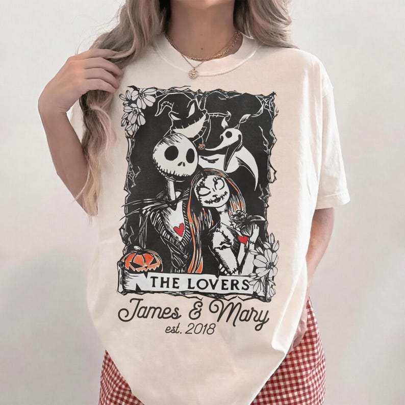 May include: Cream-colored t-shirt featuring a black and white graphic of a skeleton couple with hearts, surrounded by flowers. The text "THE LOVERS" and "James & Mary est. 2018" are printed below the image.