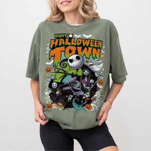 May include: Olive green t-shirt with a colorful graphic featuring characters from "The Nightmare Before Christmas." The design includes Jack Skellington, Sally, and other Halloween Town figures, with the text "Night Halloween Town."